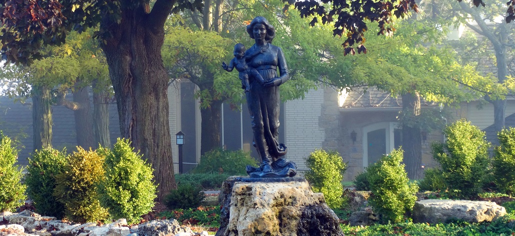 stature of Blessed Mother offering infant Jesus placed in wooded garden in front of a house